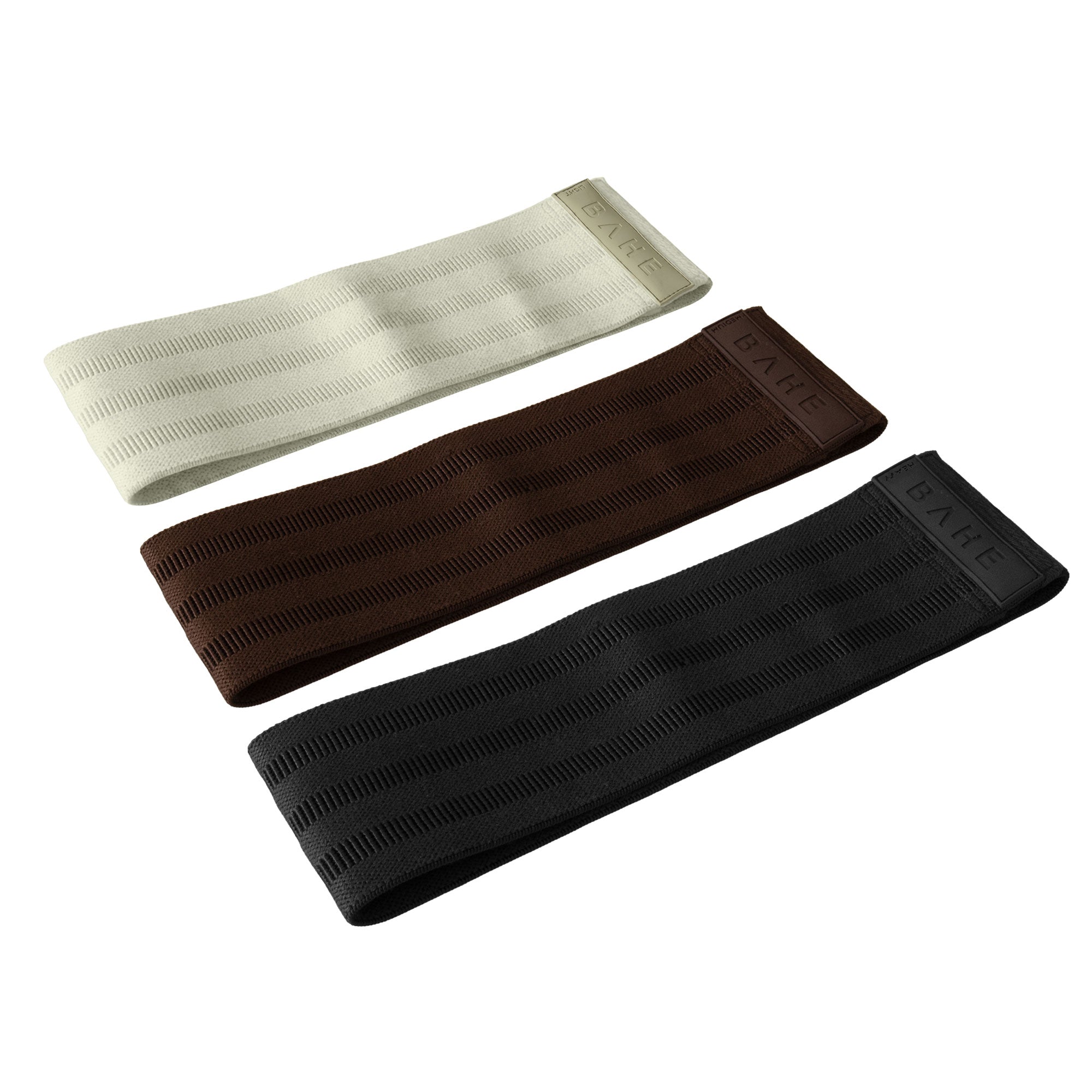 Three booty band resistance straps for pilates in cream, brown, and black, arranged diagonally on a solid white background. Each band features a ribbed texture and a small end tab with the brand name 'BAHE' embossed.