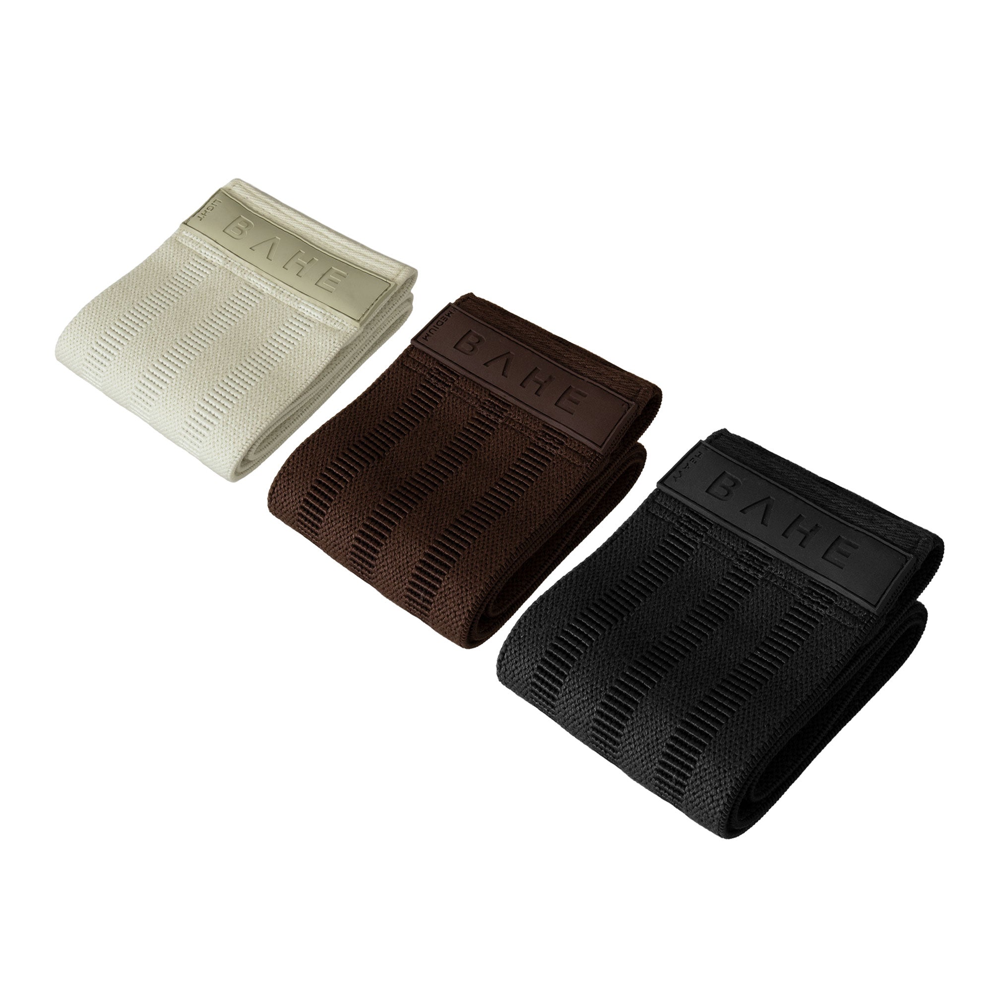 Three booty band resistance straps for pilates in cream, brown, and black, arranged diagonally on a solid white background. Each band features a ribbed texture and a small end tab with the brand name 'BAHE' embossed.